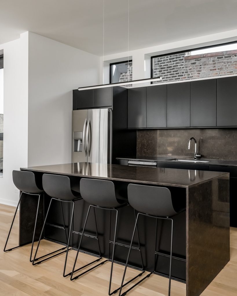 Modern kitchen with stainless steel appliances, black cabinets and dark brown granite counters and backsplash with waterfall island and black stools