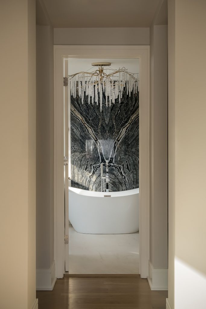 Dramatic view of primary bathroom with crystal chandelier, freestanding tub, and solid marble backsplash.